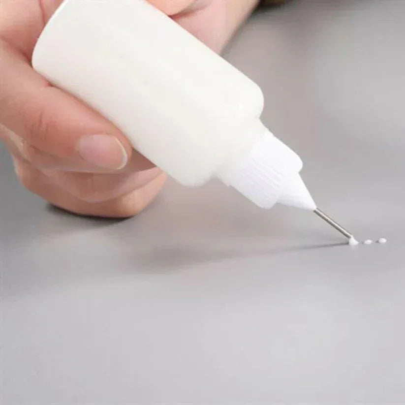 Hand holding a small white bottle with a needle-like tip, dispensing a small amount of liquid onto a gray surface.