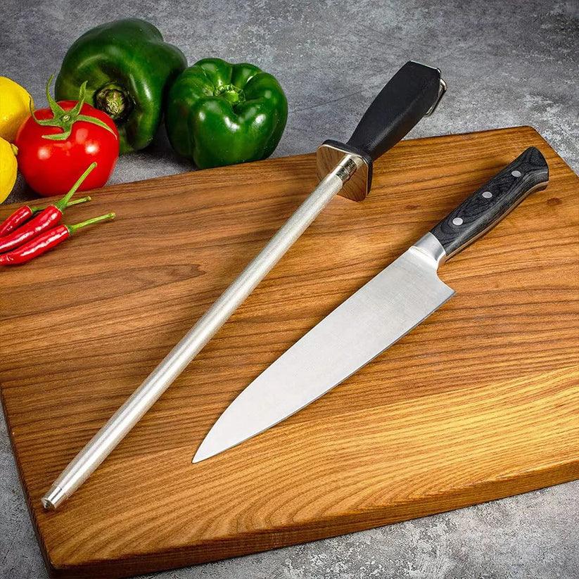Two knives on a wooden cutting board with vegetables in the background


