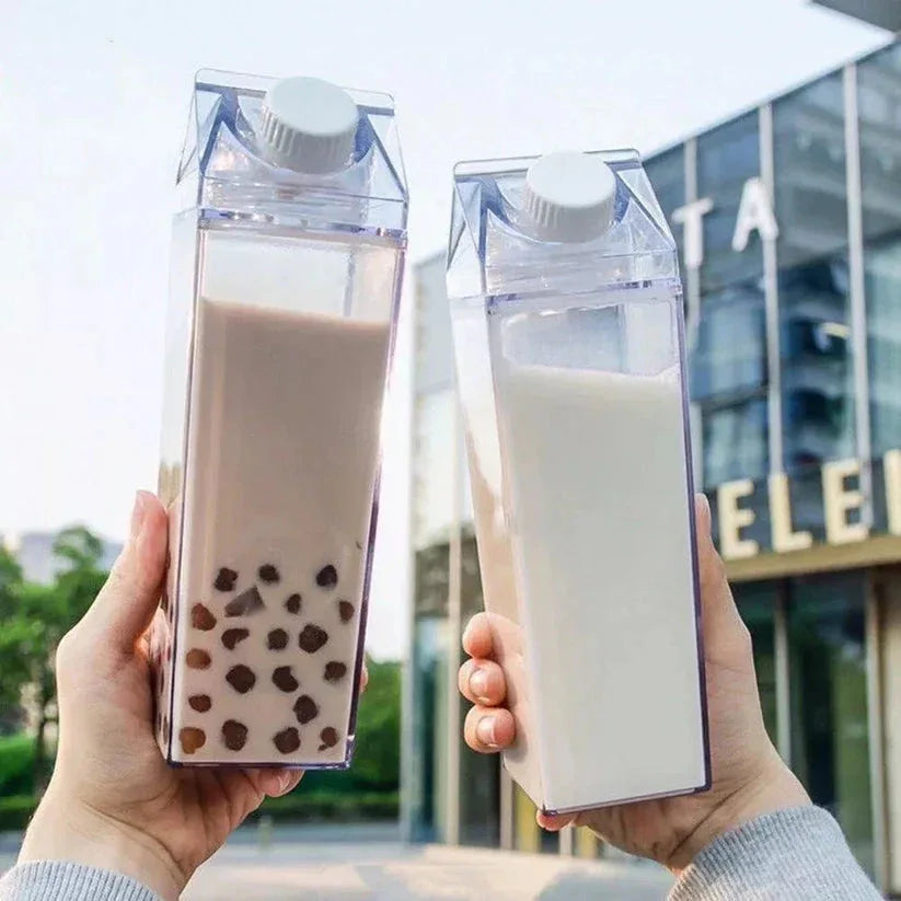 Two hands holding clear cartons of bubble tea and milk against a blurred outdoor background.

