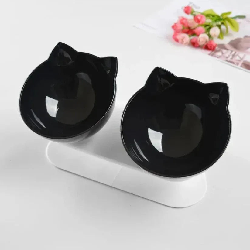 Two black cat-shaped bowls on a white stand with a magazine and flowers in the background.


