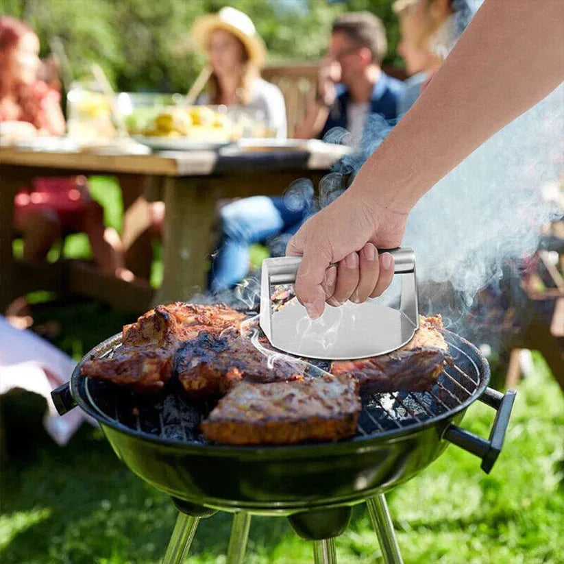 Person grilling meat on a small barbecue grill with people sitting at a table in the background.

