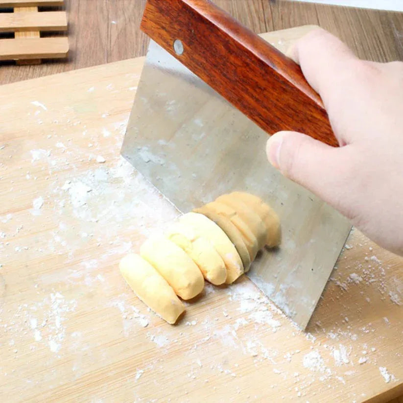 Person using a metal bench scraper to cut dough on a wooden surface.

